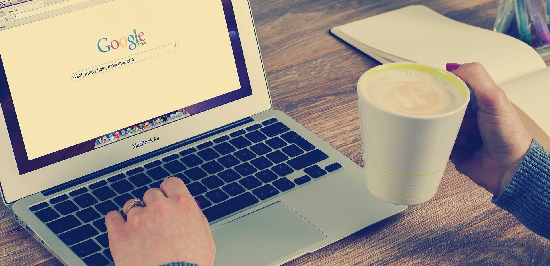 Image showing open MacBook Air with one hand on keyboard and a coffee in the other hand with open notebook on desk beside 